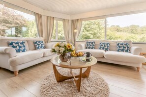 Ein modernes Wohnzimmer mit einem Glastisch, zwei weißen Sofas mit blauen Kissen und Blick ins Grüne.