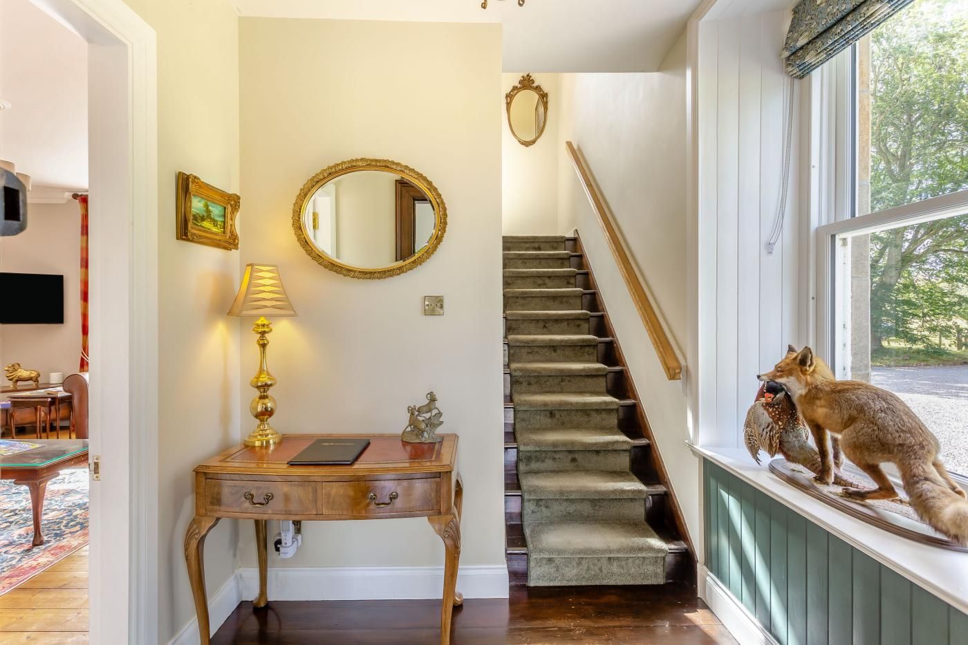 A staircase with a wooden handrail and carpeted steps, a desk with a lamp and a framed picture, and a window with a fox statue.