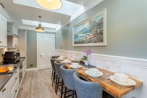 A dining area with a long wooden table set for six, a kitchen with modern appliances, and a painting on the wall. - 3 Bed in Inverarnie (oc-t45997) (Balnafoich)