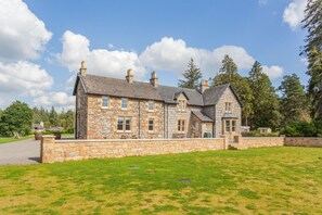 A stone house with multiple chimneys, a grassy lawn, and trees in the background. - 3 Bed in Inverarnie (oc-t45997) (Balnafoich)