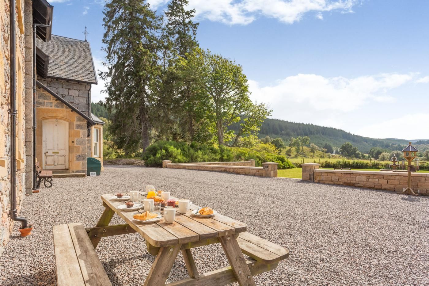 A rustic outdoor dining area with a stone building, a wooden table set with food and drinks, and a scenic view of hills and trees.