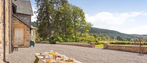A rustic outdoor dining area with a stone building, a wooden table set with food and drinks, and a scenic view of hills and trees.