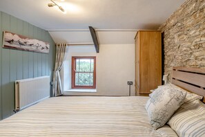 A bedroom with a bed, a window, a radiator, a wooden wardrobe, and a stone wall.