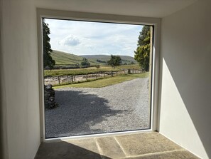 Ein Zimmer mit großem Fenster, das Blick auf eine ländliche Landschaft mit sanften Hügeln, einem Kiesweg und einer Steinmauer bietet.