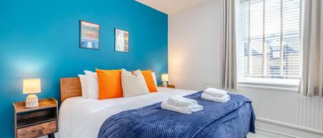 A hotel room with a bed, two bedside tables, a window with curtains, and a blue wall.