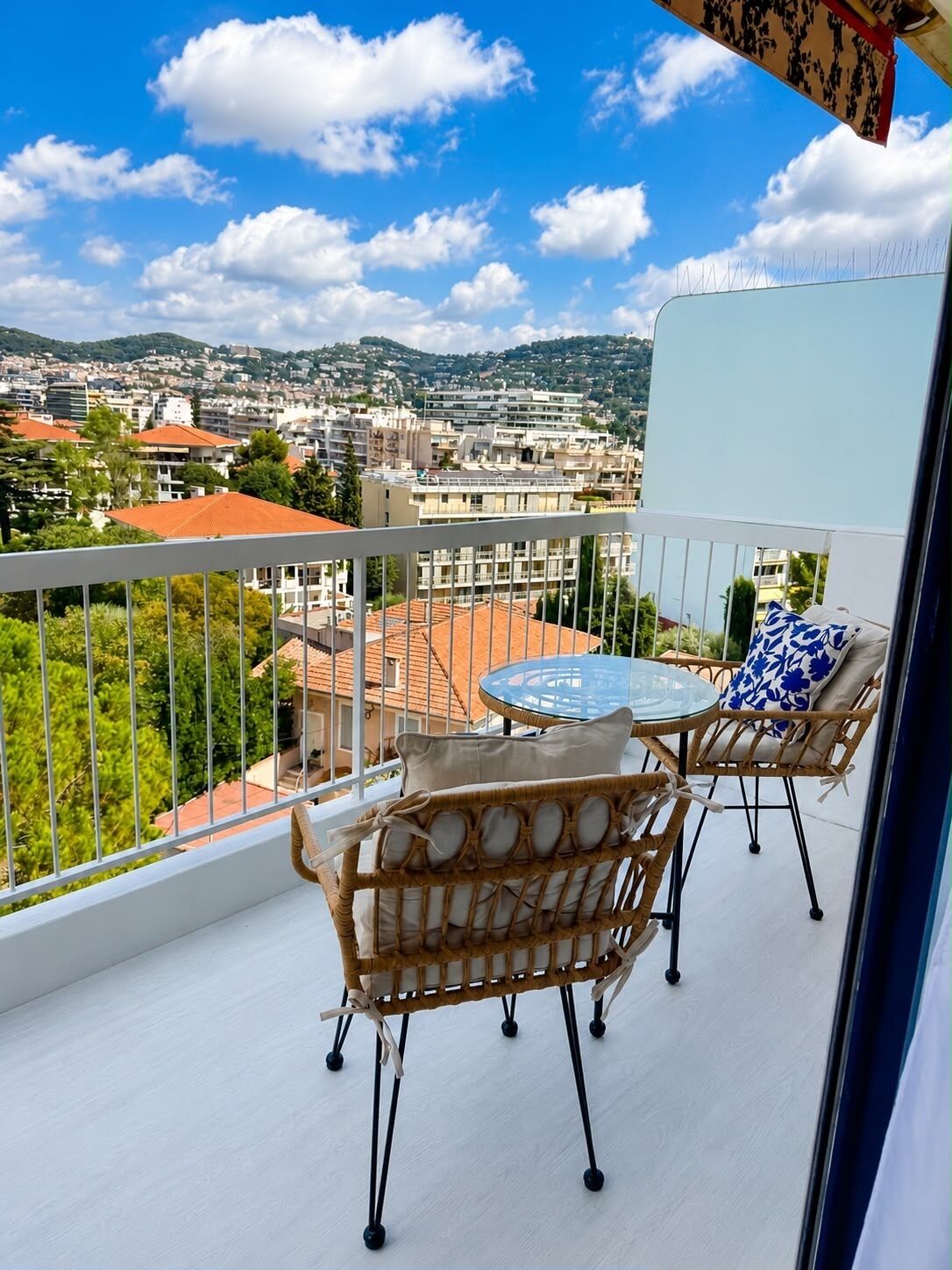 A balcony with wicker furniture and a glass table, overlooking a cityscape with buildings and greenery.