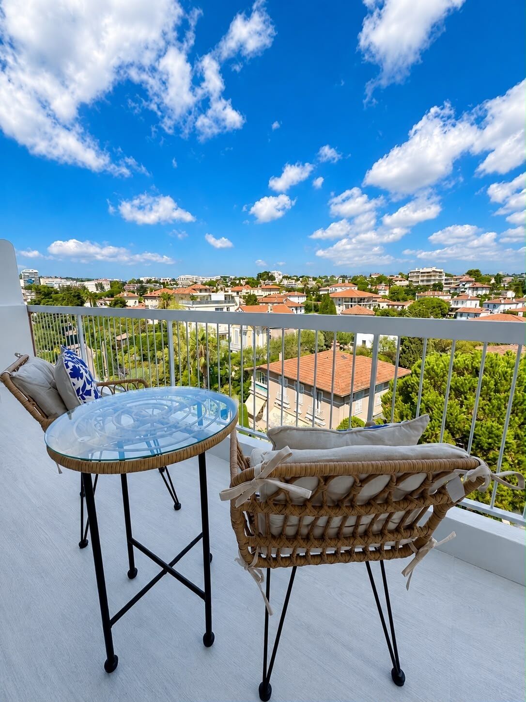 A balcony with a glass table and wicker furniture, overlooking a residential area with houses and greenery.