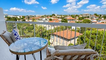 A balcony with a glass table and wicker furniture, overlooking a residential area with houses and greenery.