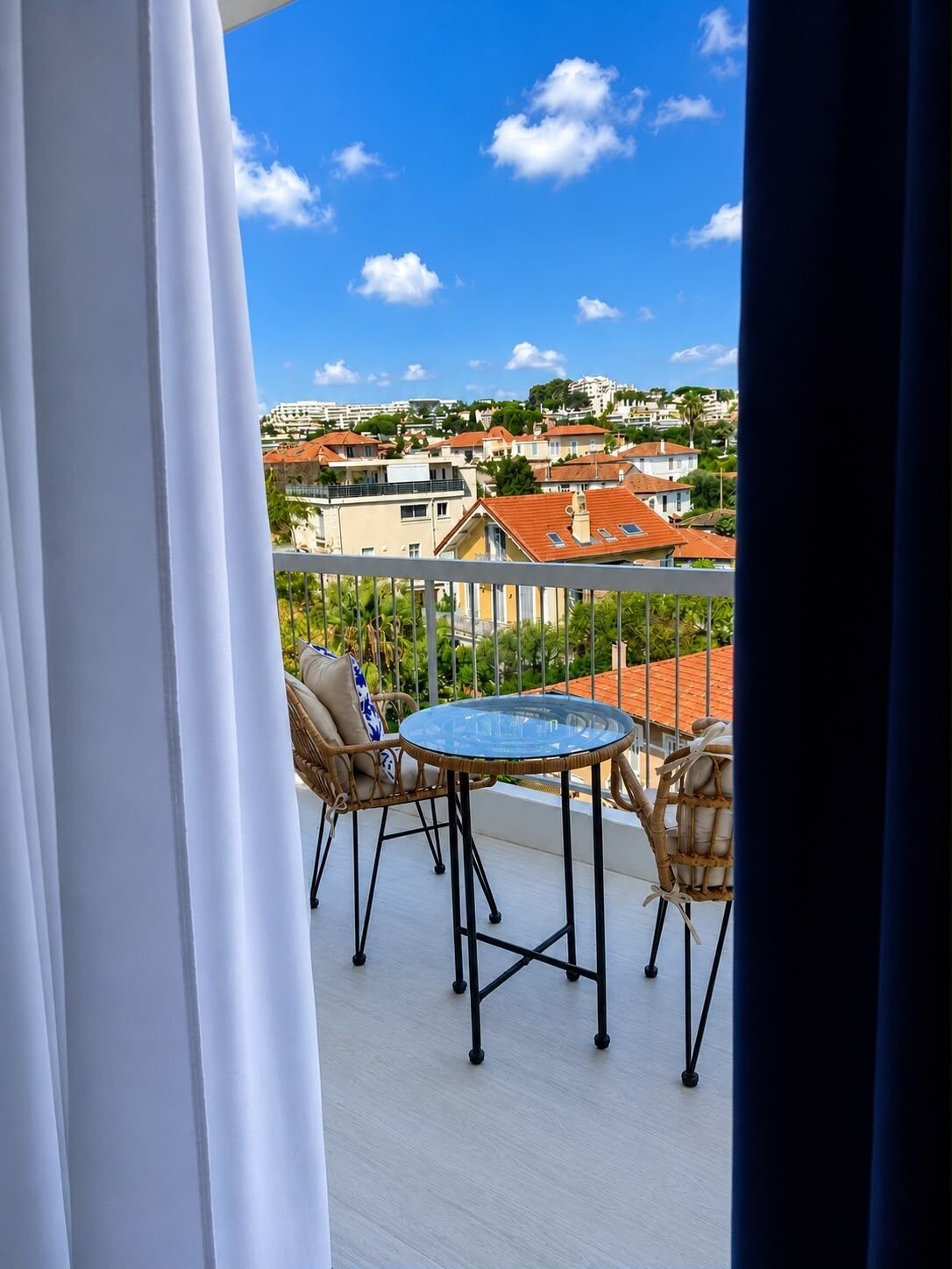 Een balkon met een ronde tafel en twee rieten stoelen, met uitzicht op een woongebied met huizen en bomen.
