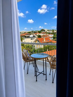Un balcon avec une table ronde et deux chaises en osier, donnant sur un quartier résidentiel avec des maisons et des arbres.