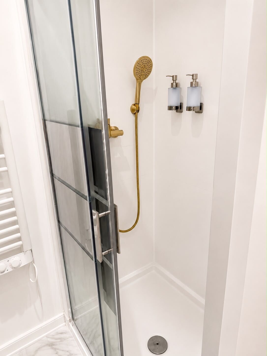 Doucheomgeving met glazen wanden, een gouden douchekop en twee zeepdispensers aan de muur.