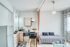 Een kleine keuken met een koelkast, magnetron en fornuis, een eethoek met stoelen, een bed met een geborduurde deken en een glazen tafel.