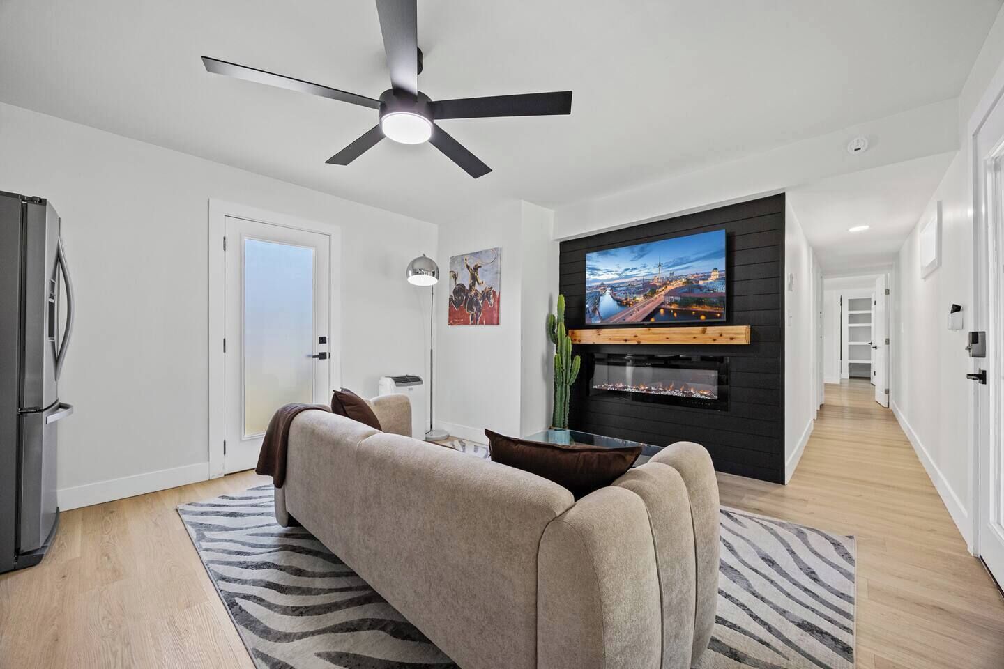 A modern living room with a ceiling fan, a flat-screen TV mounted on a dark wall, a sofa, and a cactus on a side table.
