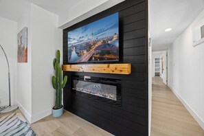 Ein modernes Wohnzimmer mit einem Flachbildfernseher an einer schwarzen Wand, einem Holzregal darunter und einem Kamin mit Glasfront.