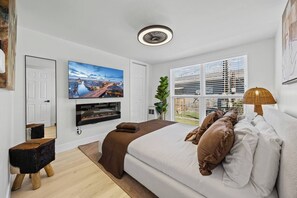 Une chambre moderne avec un grand lit, un téléviseur à écran plat fixé au-dessus, une fenêtre avec des stores et une petite table de chevet.