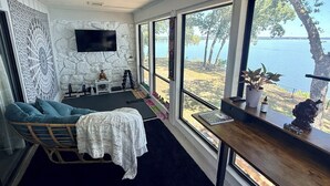 A room with a large window overlooking a body of water, a seating area with a cushioned chair, a small table, and a wall-mounted TV. - Updated Large Lakefront Townhouse with Bald Eagle Views overlooking Grand Lake. (Afton)