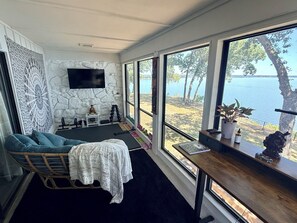 A room with a large window overlooking a body of water, a seating area with a cushioned chair, a small table, and a wall-mounted TV. - Updated Large Lakefront Townhouse with Bald Eagle Views overlooking Grand Lake. (Afton)