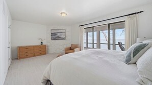 A bedroom with a large bed, a dresser, a chair, and a view of the outdoors through a window. - Updated Large Lakefront Townhouse with Bald Eagle Views overlooking Grand Lake. (Afton)