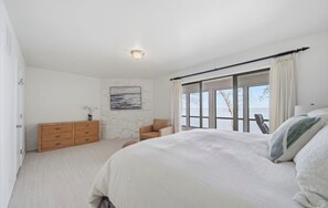 A bedroom with a large bed, a dresser, a chair, and a view of the outdoors through a window. - Updated Large Lakefront Townhouse with Bald Eagle Views overlooking Grand Lake. (Afton)