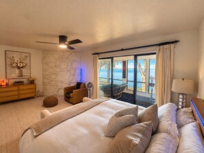 A bedroom with a large bed, a ceiling fan, a stone wall, a view of the sea, a wooden dresser, and a chair by the window.