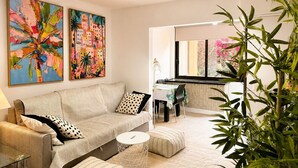 A modern living room with a sofa, ottoman, and a dining area with a table and chairs. There are two large paintings on the wall and a plant in the foreground. - Castle Comfort (Los Cristianos)