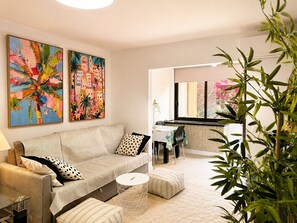 A modern living room with a sofa, ottoman, and a dining area with a table and chairs. There are two large paintings on the wall and a plant in the foreground.