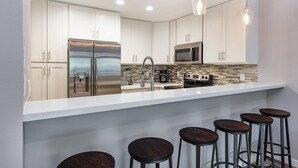 A modern kitchen with stainless steel appliances, a white countertop, and bar stools. - Tropical Garden Vibes Maui (Kihei)