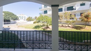 A covered outdoor area with a paved floor, a metal railing, and a view of a building with multiple balconies and trees. - Casa das Conchas T2 by YHA (Conceiçao de tavira)