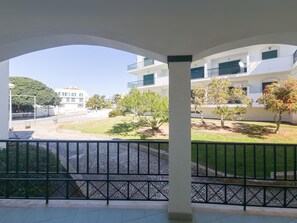 Uma área externa coberta com piso pavimentado, guarda-corpo de metal e vista para um edifício com várias varandas e árvores.