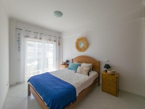 A bedroom with a bed, a nightstand, a wall-mounted mirror, and a window with sheer curtains. - Casa das Conchas T2 by YHA (Conceiçao de tavira)