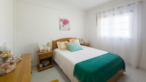 A bedroom with a bed, nightstands, a lamp, a decorative wall piece, and a window with curtains. - Casa das Conchas T2 by YHA (Conceiçao de tavira)