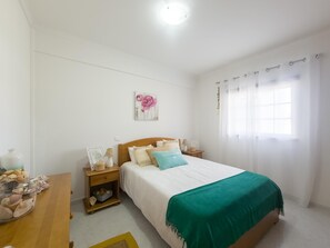 Quarto com cama, mesas de cabeceira, luminária, um objeto decorativo na parede e uma janela com cortinas.