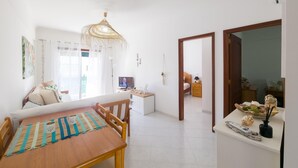 A bright living room with a wooden dining table, a sofa, and a TV. There is a hallway leading to another room. - Casa das Conchas T2 by YHA (Conceiçao de tavira)