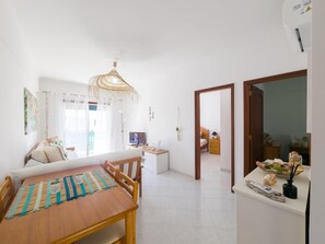 Uma sala de estar luminosa com uma mesa de jantar de madeira, um sofá e uma televisão. Há um corredor que dá para outro quarto.