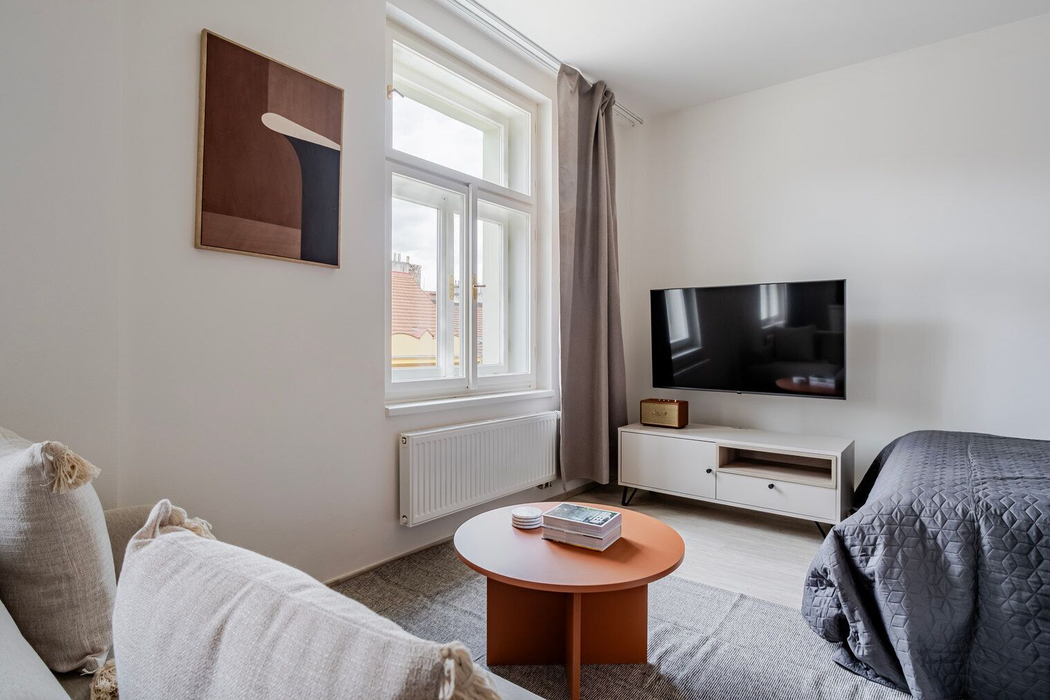 Una habitación de hotel con cama, televisión, una mesita con libros, ventana con cortinas y un radiador.