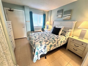 Um quarto com uma cama, mesas de cabeceira, um criado-mudo, uma janela com cortinas e uma porta.