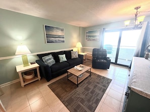 Uma sala de estar com sofá, poltronas, uma mesa de centro e vista para o mar através de uma grande janela.