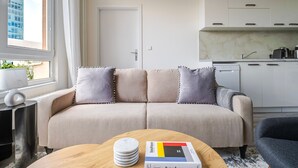 A modern living room with a sofa, a coffee table with a book, and a kitchen area with cabinets and a window. - Blueground | Nusle, city view, nr Vysehrad (PRG-108) (Prague)