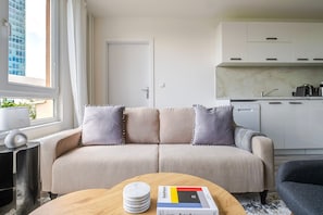 Ein modernes Wohnzimmer mit einer Couch, einem Couchtisch mit einem Buch und einer Küchenzeile mit Schränken und einem Fenster.