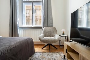 Une chambre d’hôtel avec un lit, une chaise, une télévision, une petite table avec une lampe et une fenêtre avec des rideaux.