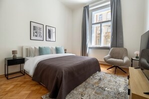 Quarto com cama, cadeira, mesa de cabeceira, abajur e janela com cortinas.