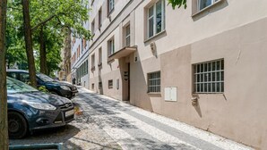 Kapea mukulakivikatu, jonka varrella on parkkeerattuja autoja ja puita, johtaa rakennukseen, jonka julkisivu on tasainen ja jossa on useita ikkunoita.