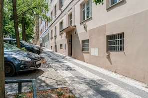 A narrow cobblestone street lined with parked cars and trees, leading to a building with a flat facade and multiple windows. - Blueground | Vinohrady, view, nr Vysehrad (PRG-109) (Prague)