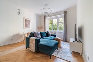 A modern living room with a sofa, a side table, a lamp, a TV, and a large window with curtains. - Blueground | Vinohrady, view, nr Vysehrad (PRG-109) (Prague)