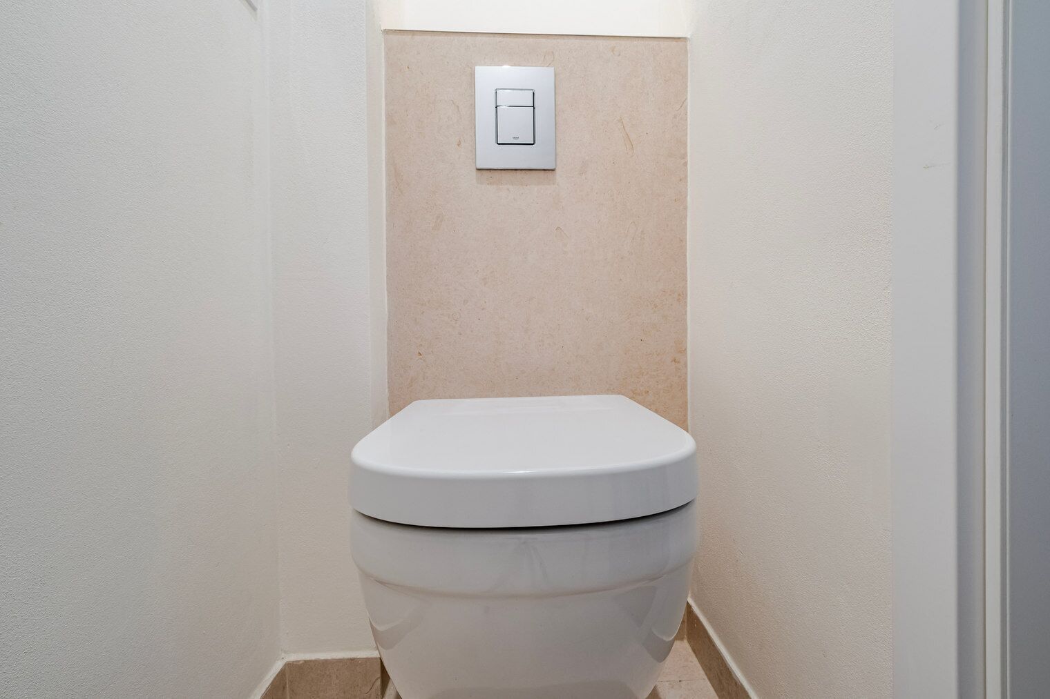 Des toilettes blanches dans une petite salle de bain, avec un mur de couleur beige et un interrupteur situé au-dessus.