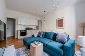 A modern living room with a teal sofa, a small wooden side table, and a kitchen in the background. - Blueground | Vinohrady, view, nr Vysehrad (PRG-109) (Prague)