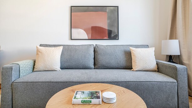 Ein graues Sofa mit gemusterten Kissen, ein gerahmtes abstraktes Gemälde, eine Lampe und ein Tisch mit einem Buch und Untersetzern.