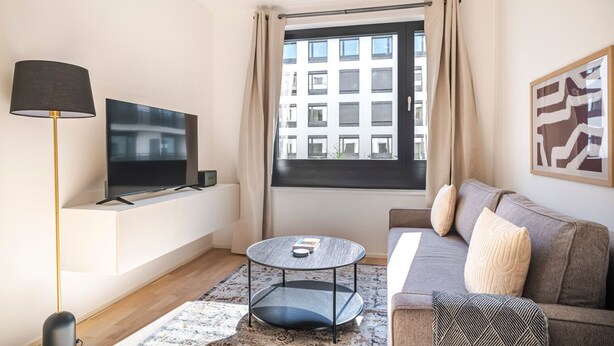 Een moderne woonkamer met een flatscreen televisie, een ronde salontafel, een bank met kussens, een vloerlamp en een raam met gordijnen.