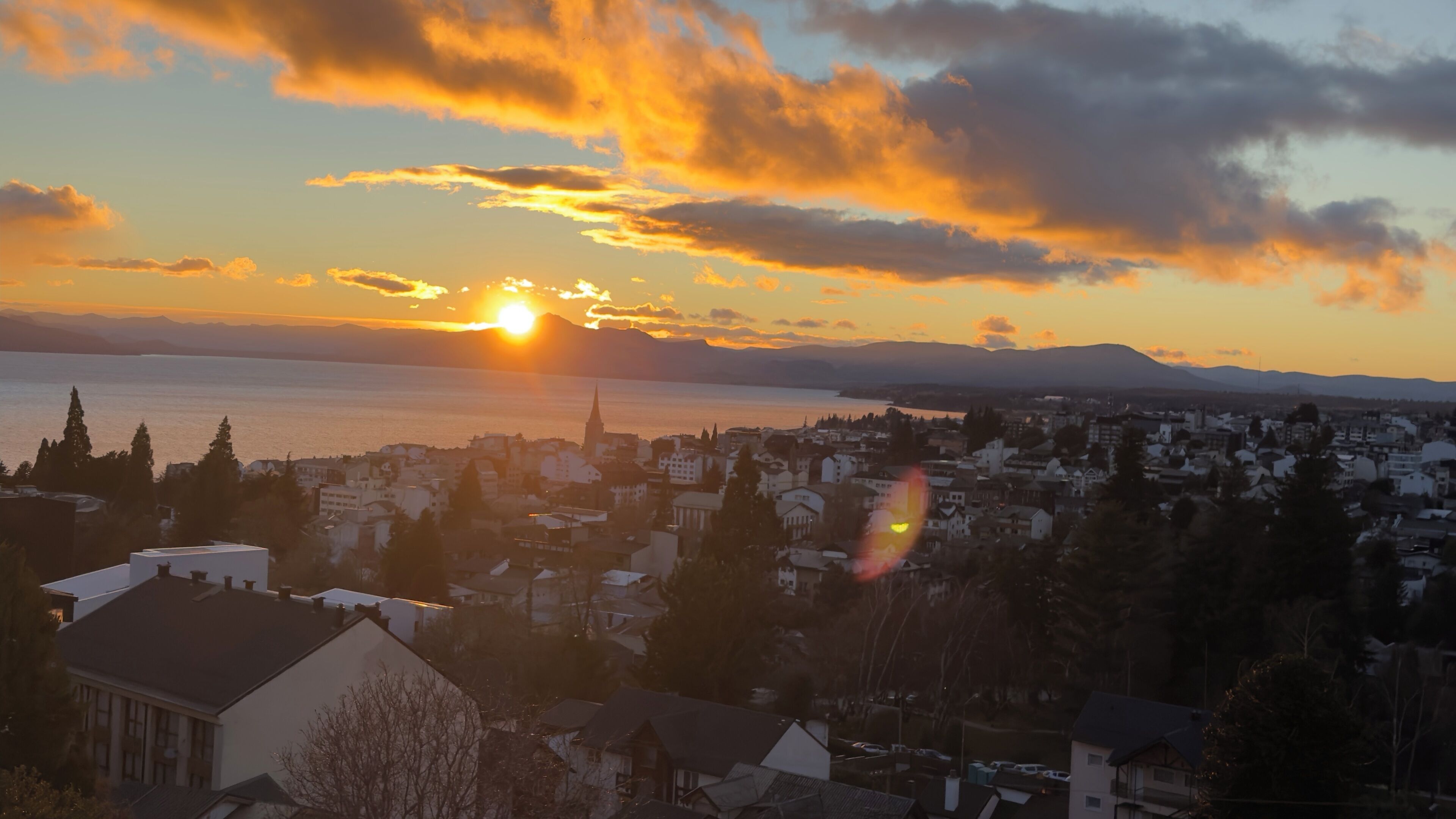 A cityscape at sunset with a body of water in the foreground and mountains in the distance.