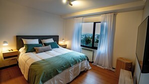 A bedroom with a large bed, bedside lamps, a window with curtains, and a view of the city. - El Coral - Lake View Apartment · Great Location in San Carlos de Bariloche (San Carlos de Bariloche)